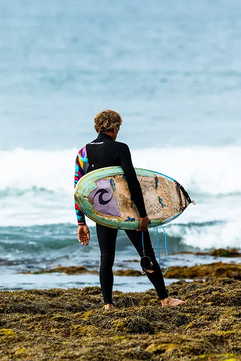 Tom Curren standing on the rocks with a surfboard under his arm